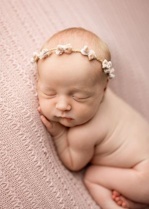 Sleeping newborn baby with delicate white floral headpiece rests peacefully on soft pink textured blanket.