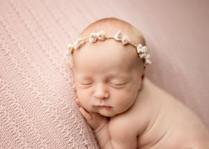 Peaceful sleeping newborn baby wearing delicate floral headband on soft pink textured blanket.