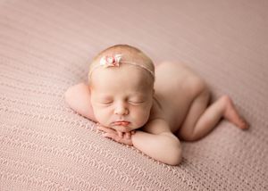 Peaceful sleeping newborn baby curled up on soft pink textured blanket wearing delicate bow headband.
