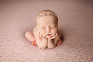 Newborn baby sleeping in frog pose on pink blanket wearing delicate gold headband during professional photo session.