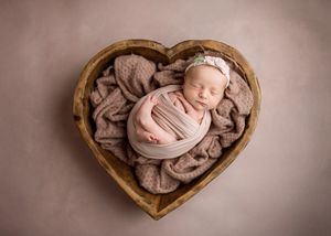 A sleeping newborn swaddled in soft fabric lies peacefully in a wooden heart-shaped bowl filled with textured blankets.