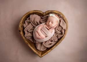 Newborn baby wrapped in soft pink swaddle sleeping peacefully in wooden heart-shaped bowl with beige blanket.
