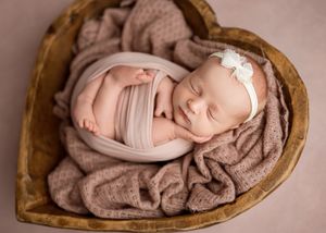 A peaceful sleeping newborn wrapped in soft pink fabric lies in a heart-shaped wooden bowl with taupe blanket.