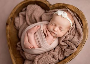 A peaceful newborn baby wrapped in soft pink fabric sleeps in a heart-shaped wooden bowl with brown blanket.