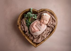 Newborn baby wrapped in pink fabric sleeping peacefully in wooden heart-shaped bowl with greenery accent.