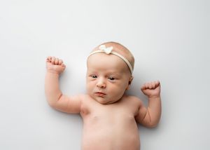 A peaceful baby lying on a white background wearing a neutral headband with arms raised.