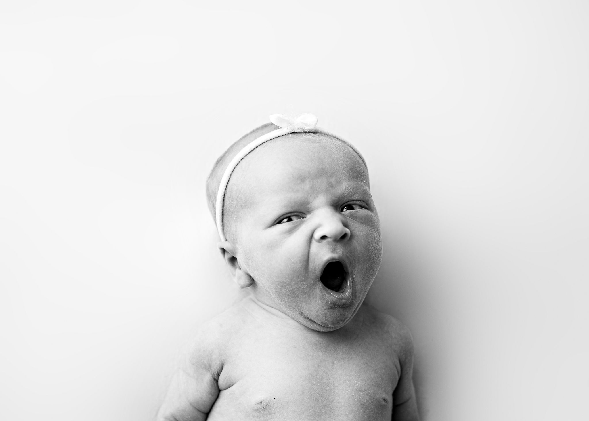 A black and white portrait of a yawning baby with a small white cap against a white background.