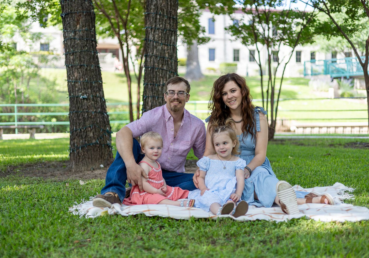 Family enjoying a summer picnic together while sitting on a blanket under shady trees in a park setting.