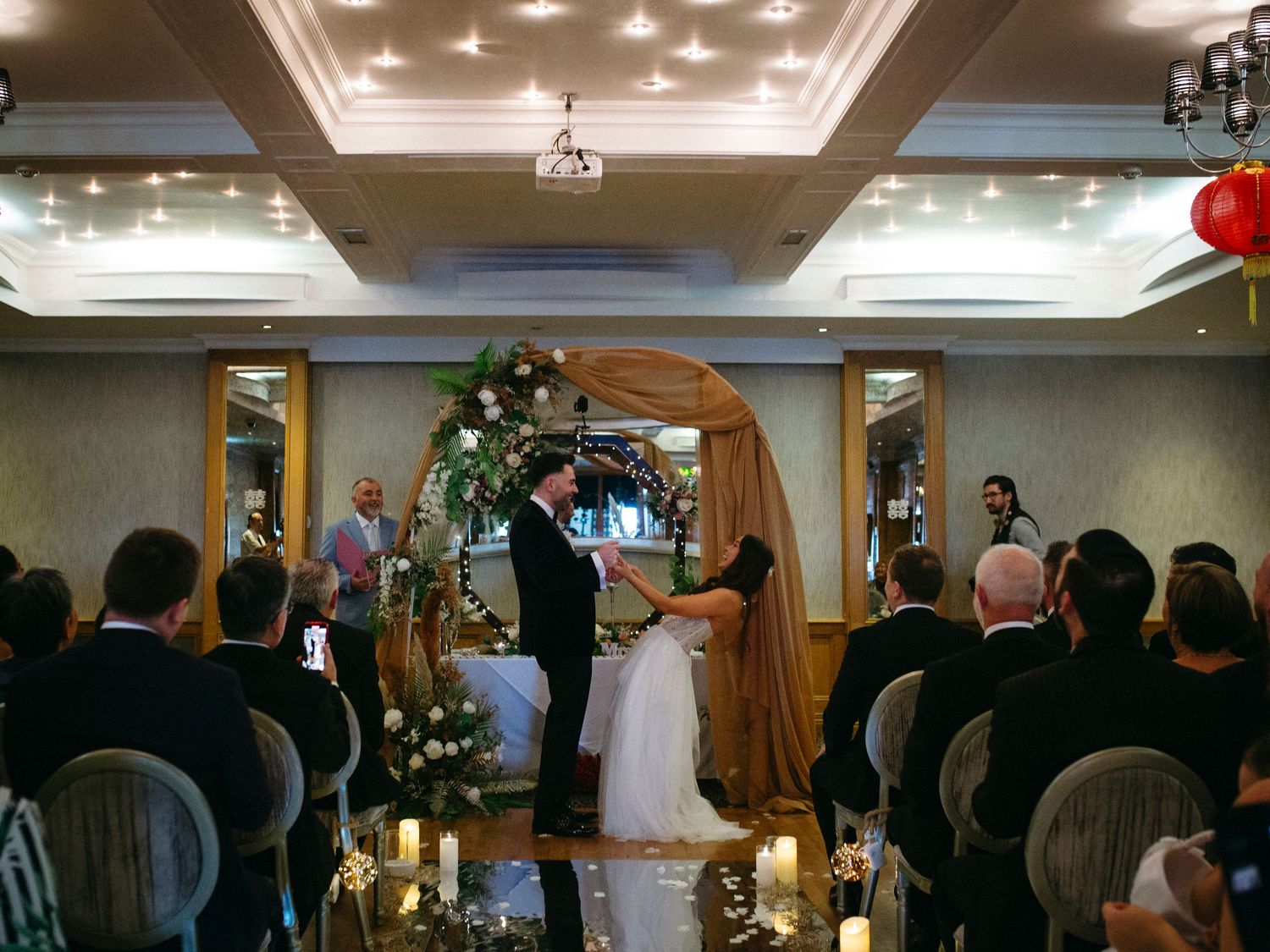Wedding ceremony in an elegant ballroom with draped arch, floral arrangements and candle-lit aisle decorations.