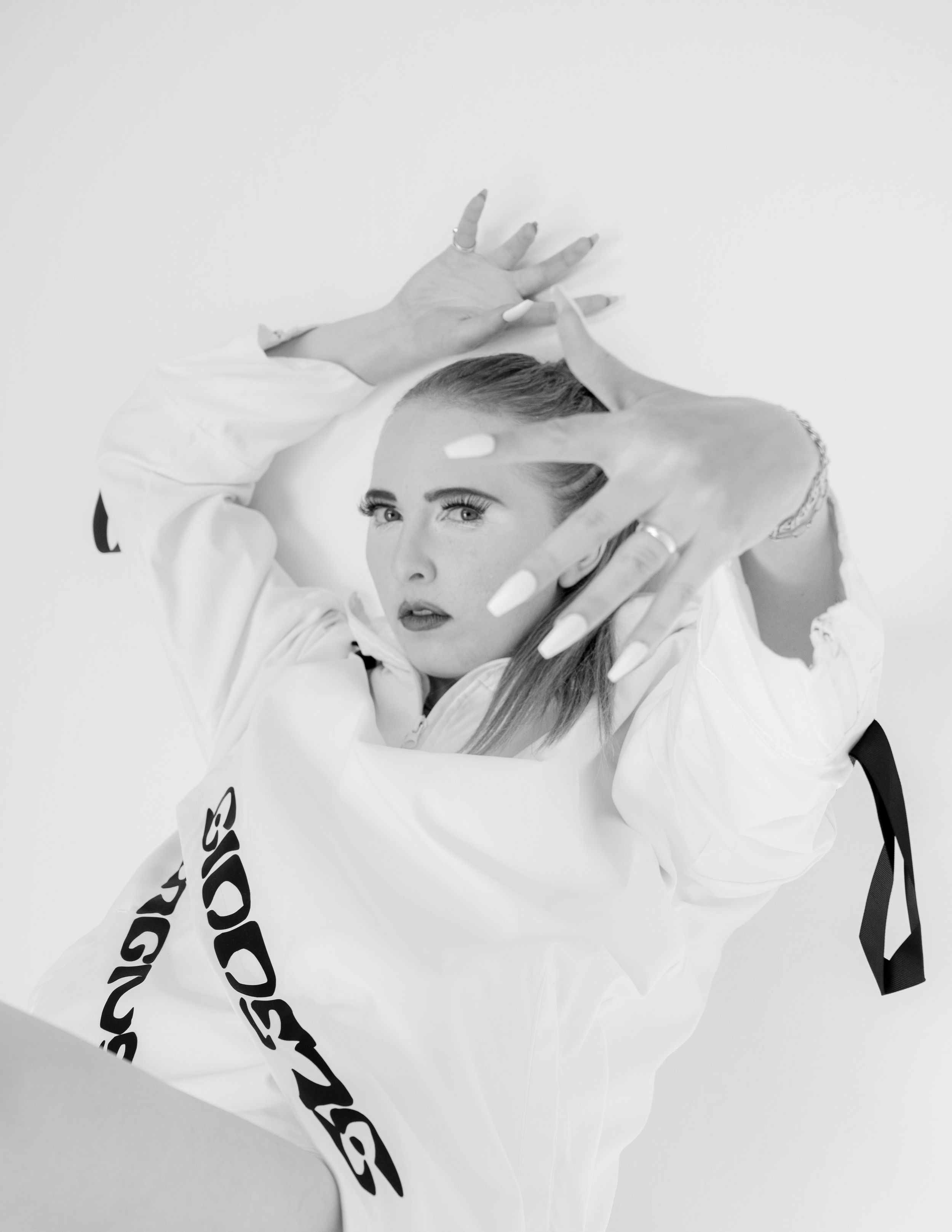Artistic black and white photo of a person posing in a white outfit with dramatic hand gestures against plain backdrop.