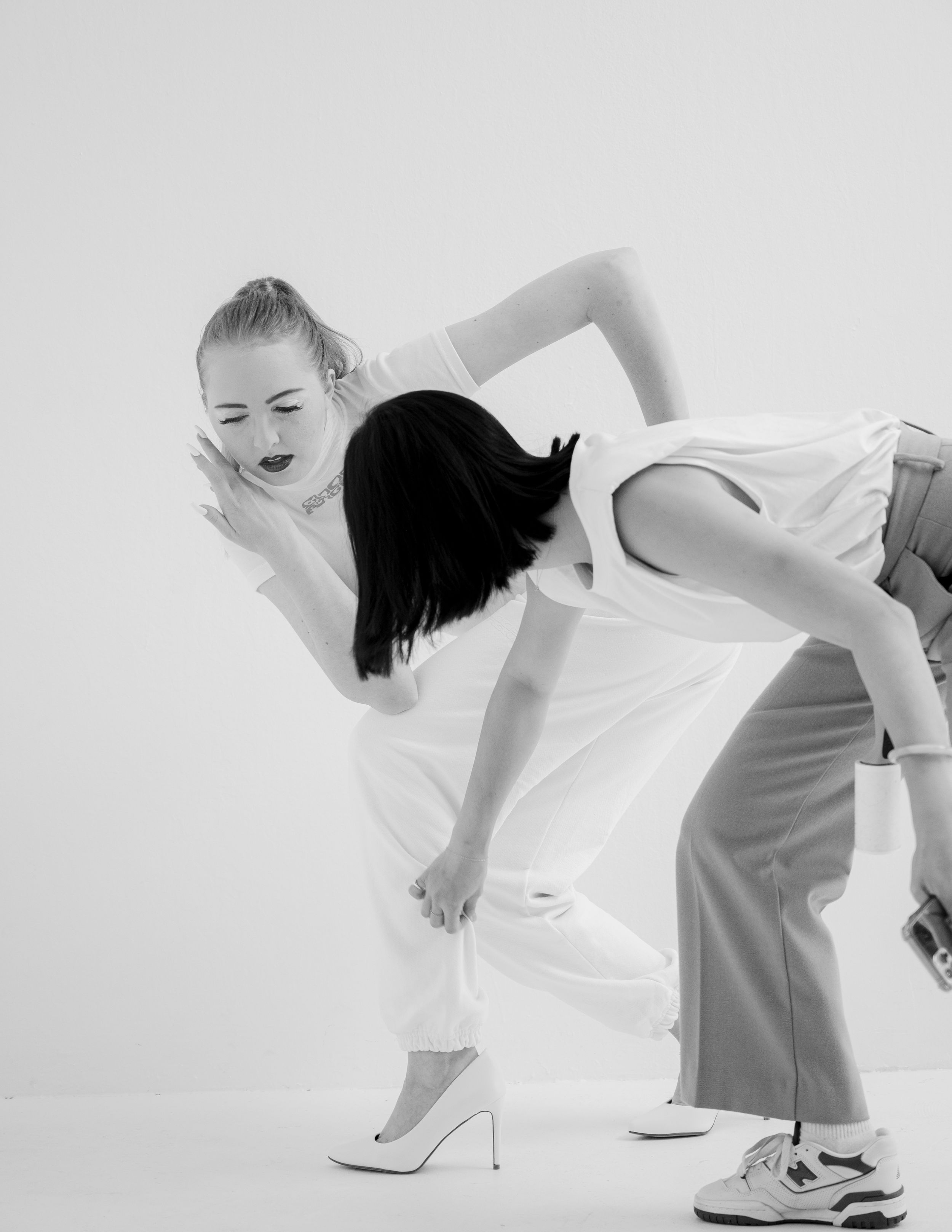 A dynamic dance pose in black and white shows two dancers interacting, one bending forward in heels and white outfit.