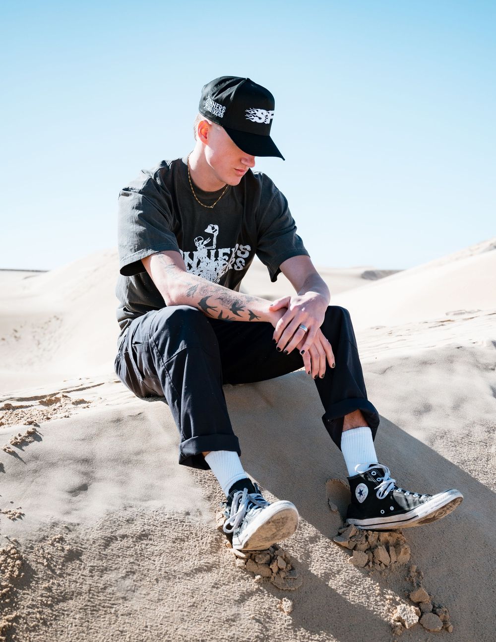 Person wearing a black hat and clothing sits on sandy terrain against a blue sky, creating a minimalist desert scene.