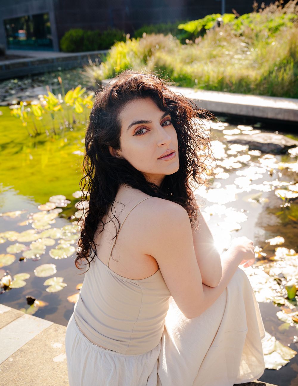 Woman wearing beige top poses thoughtfully by pond on sunny day with fallen leaves floating on water's surface.