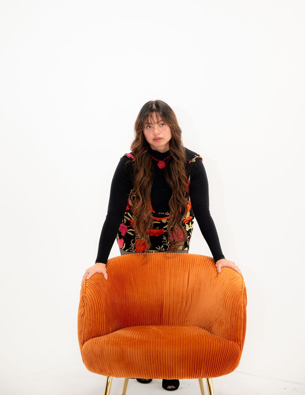 A stylishly dressed woman in a black turtleneck and patterned vest stands behind a retro orange corduroy armchair.