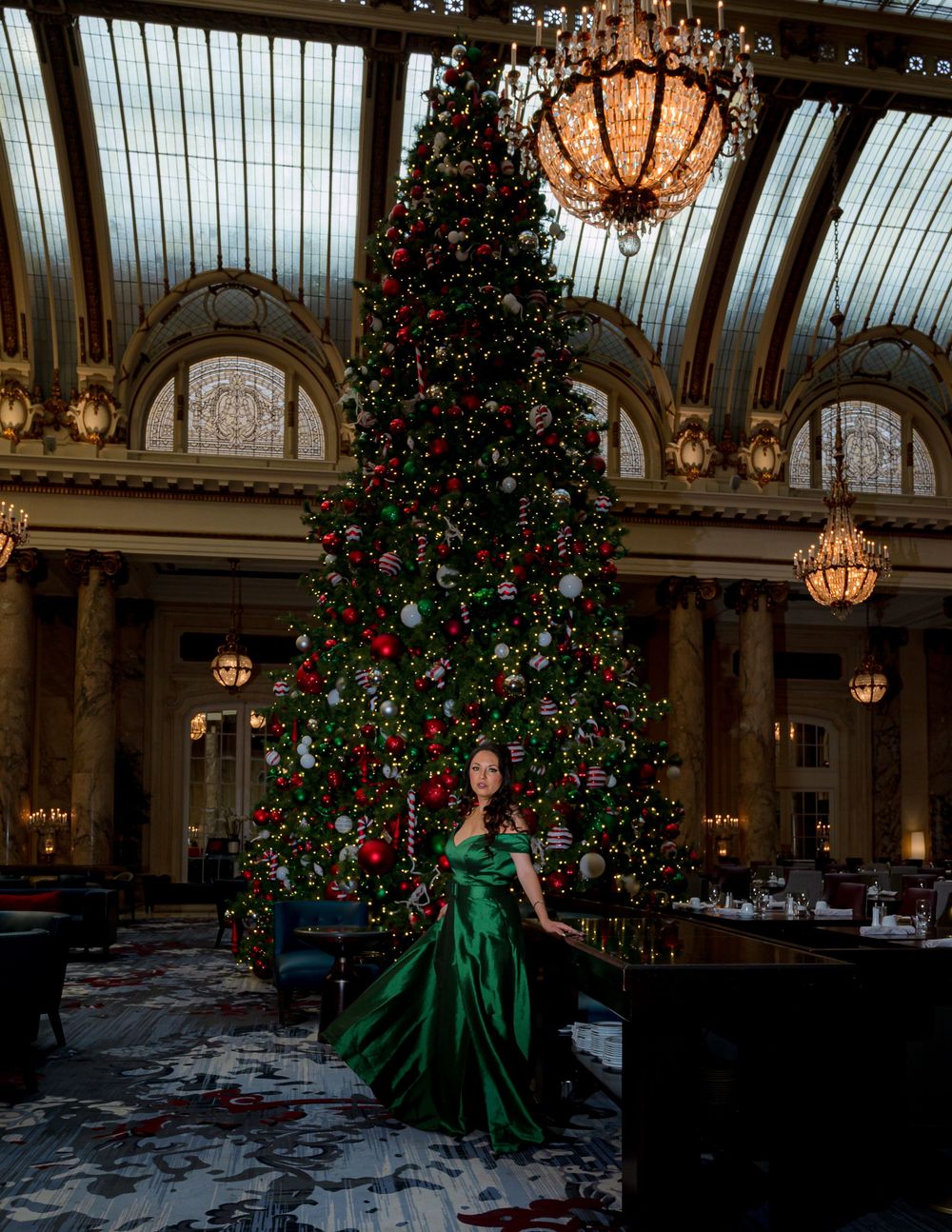 Elegant figure in emerald green gown poses dramatically in ornate ballroom with crystal chandeliers and Christmas trees.