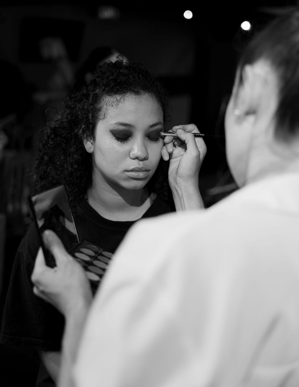 Black and white image showing someone getting makeup applied by a makeup artist in a dark setting.