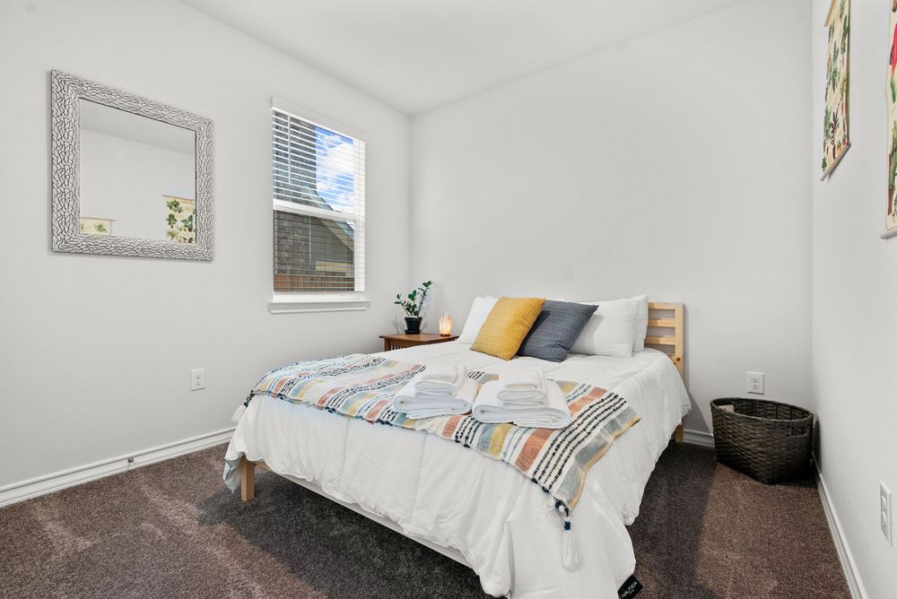 Modern bedroom with white walls, a window, gray framed mirror, and bed with colorful throw pillows on dark carpet.