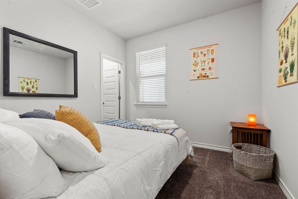 A modern bedroom with white walls features a mirror, bed with white bedding and orange accent pillow, and wall art decor.