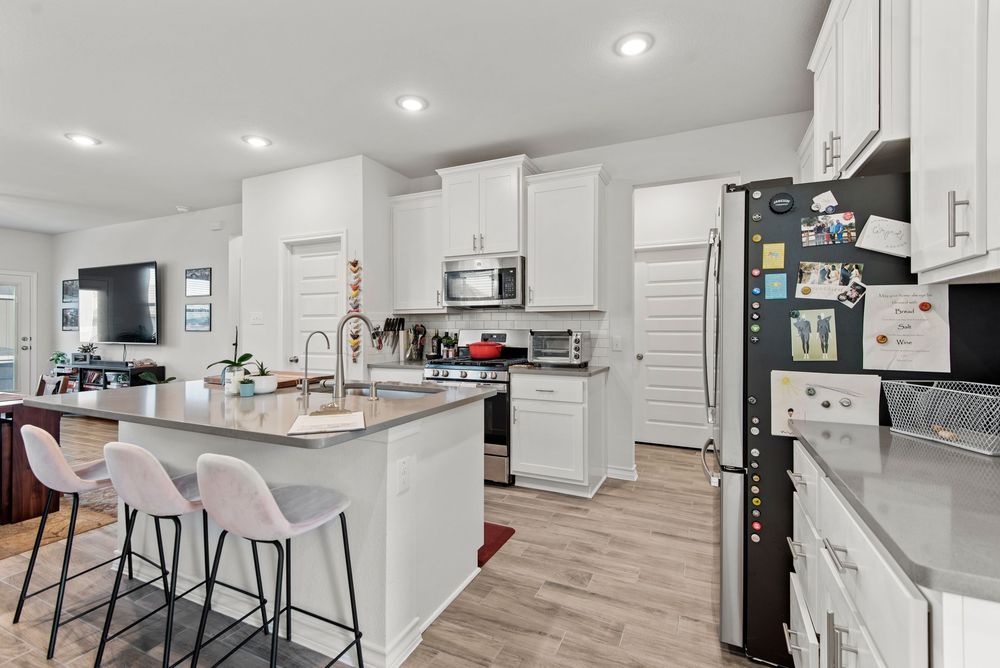 Modern white kitchen featuring quartz countertops, bar seating, stainless appliances and grey wood-style flooring.