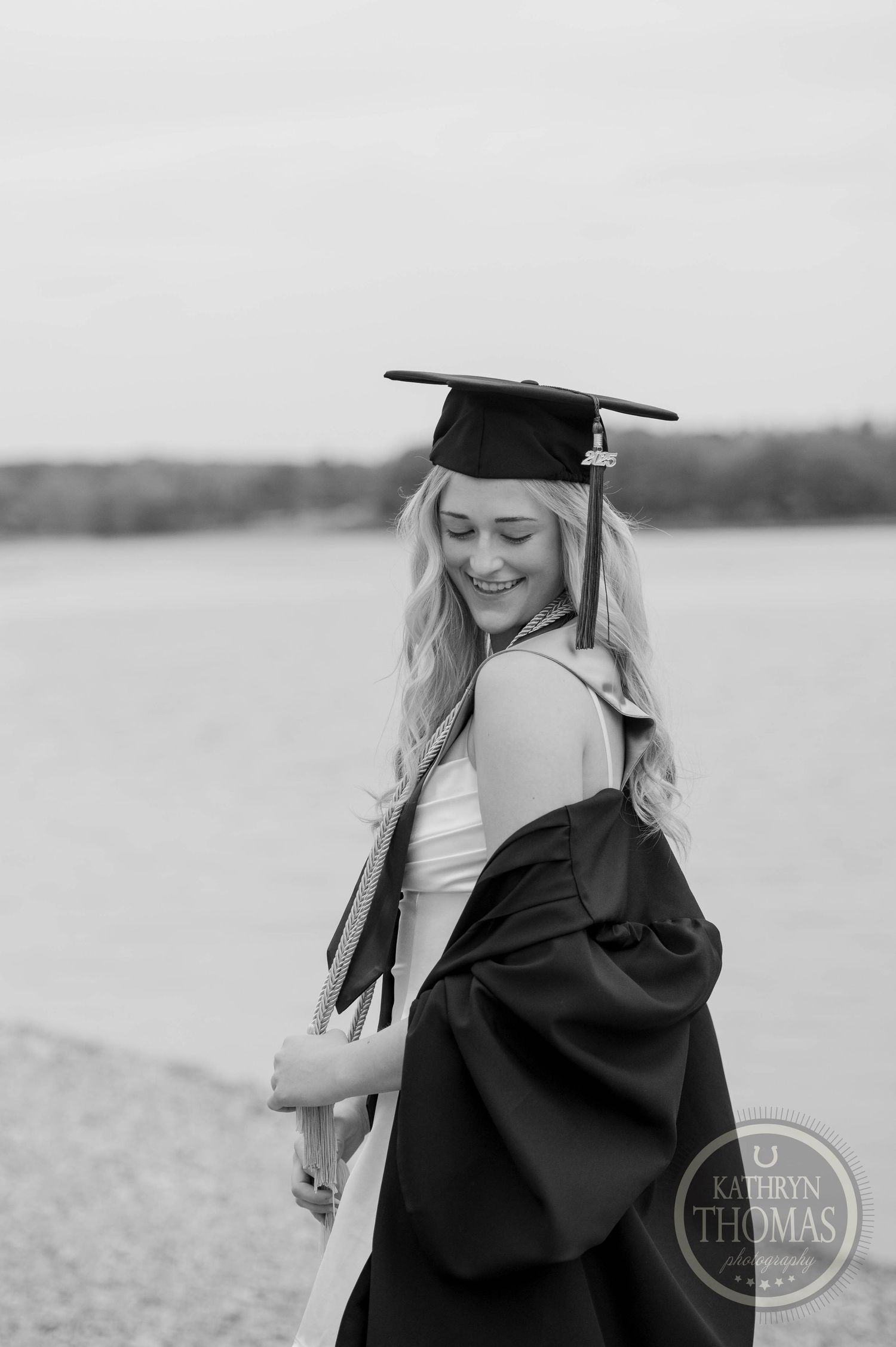 Black and white graduation portrait of a graduate in cap and gown smiling by a lake.