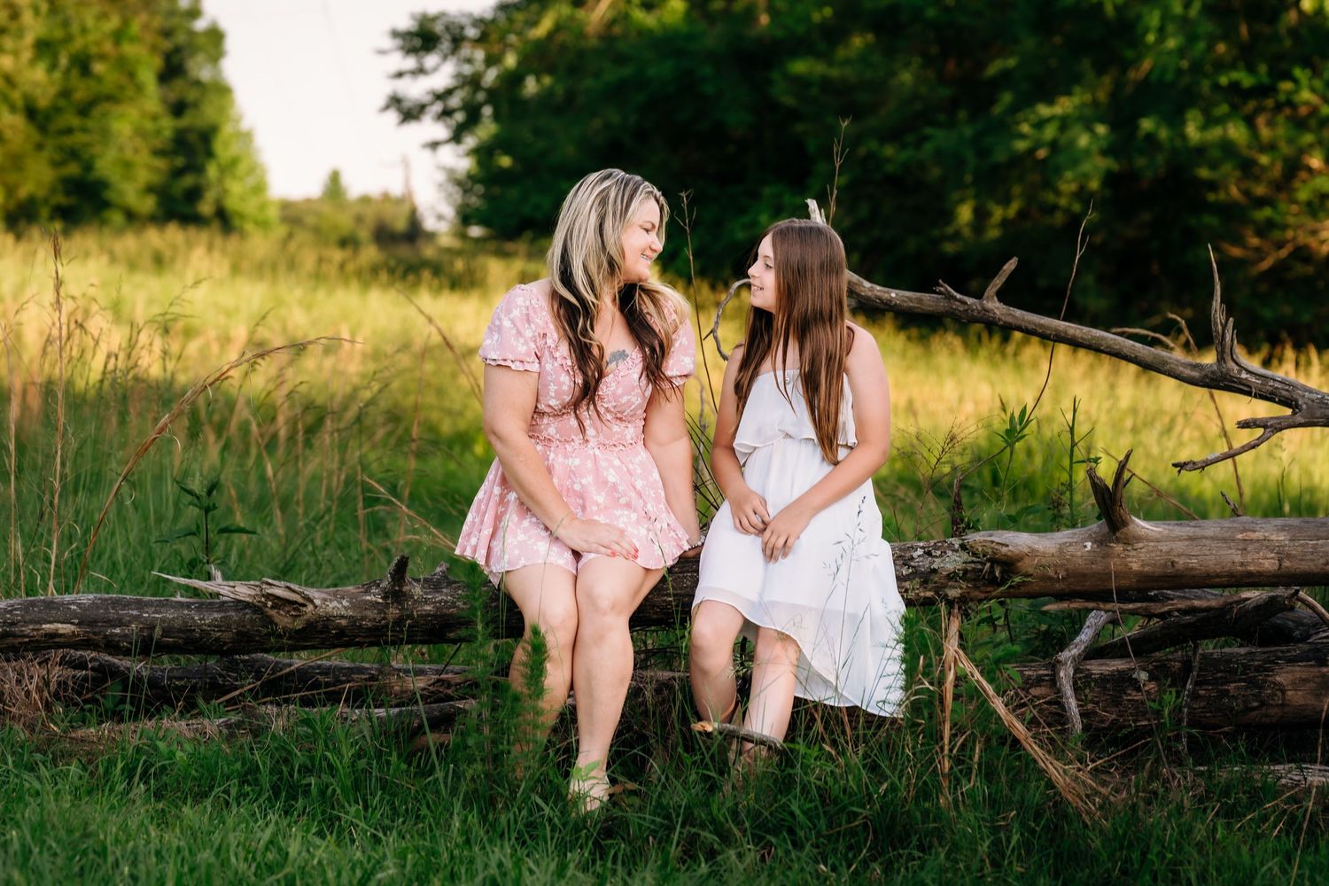 Two people in white dresses share a moment while sitting on fallen logs in a sunlit summer meadow.