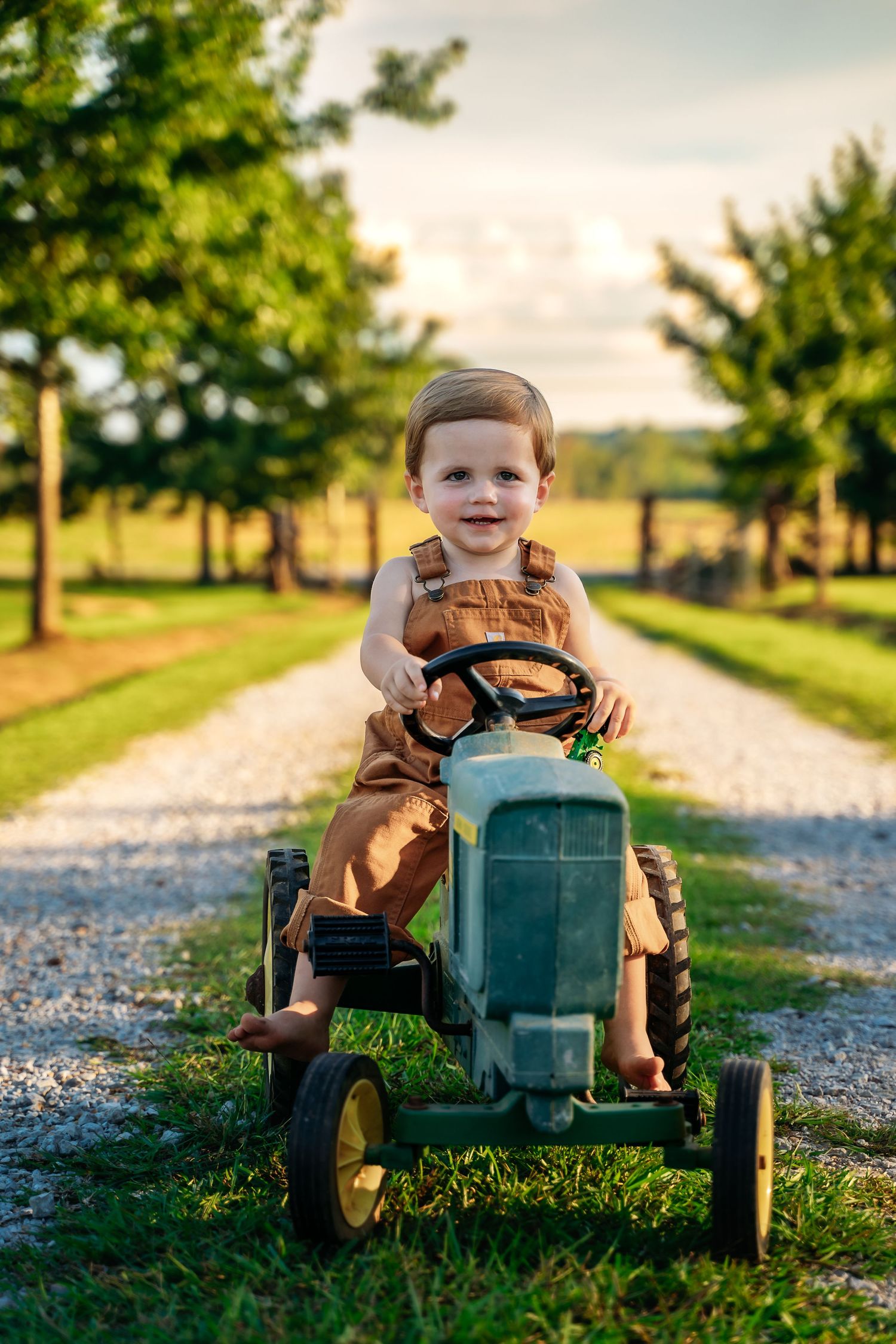 A child rides a green toy tractor along a gravel path on a sunny day as trees line the rural road.