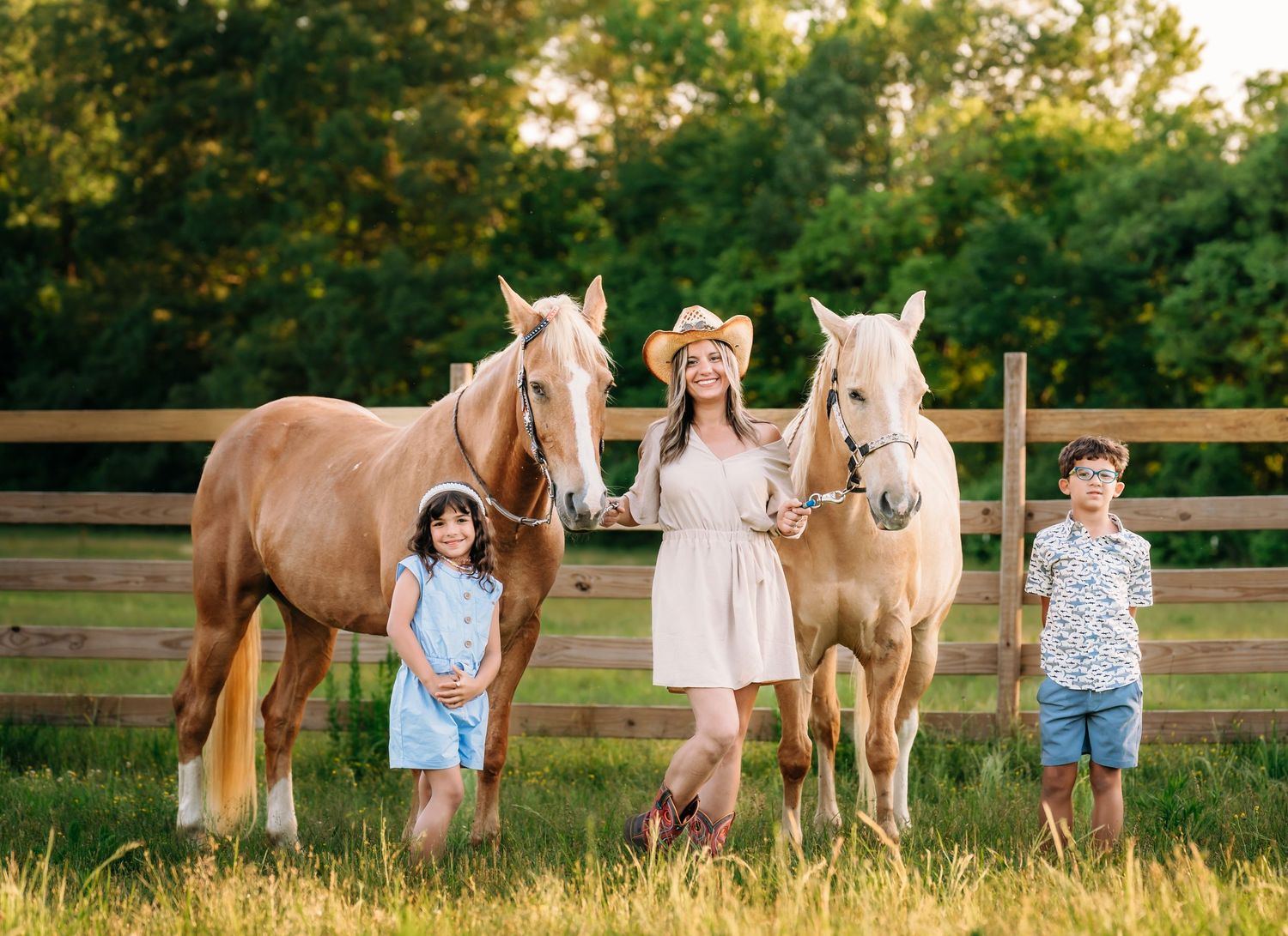 Three children pose with two palomino horses in front of a wooden fence during golden hour on a summer evening.