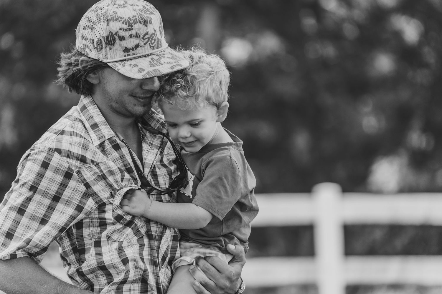 A tender black and white moment captured between a person in a plaid shirt and hat embracing a young child outdoors.
