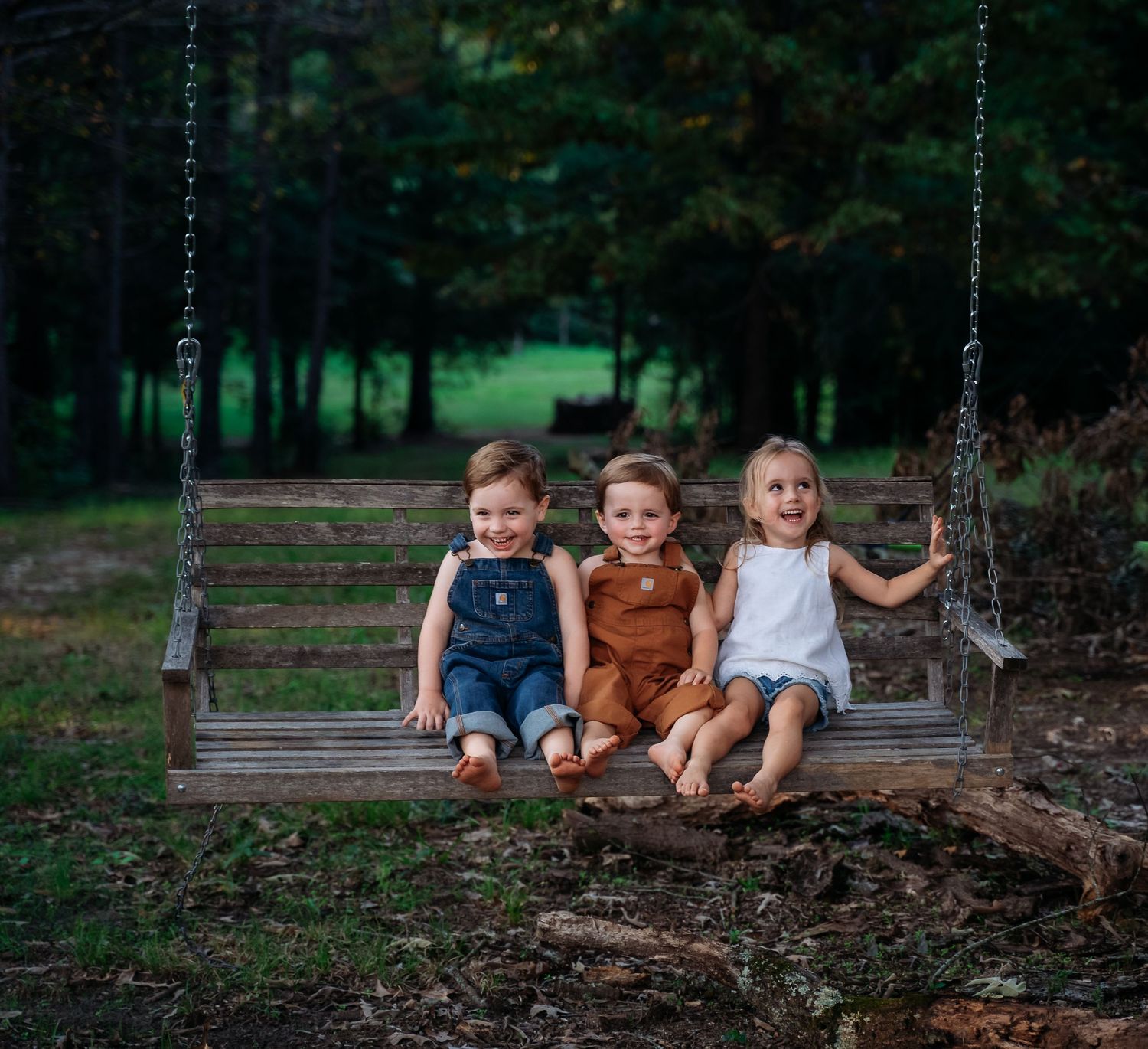 Three small children sit together smiling on a rustic wooden swing in a serene forest setting.