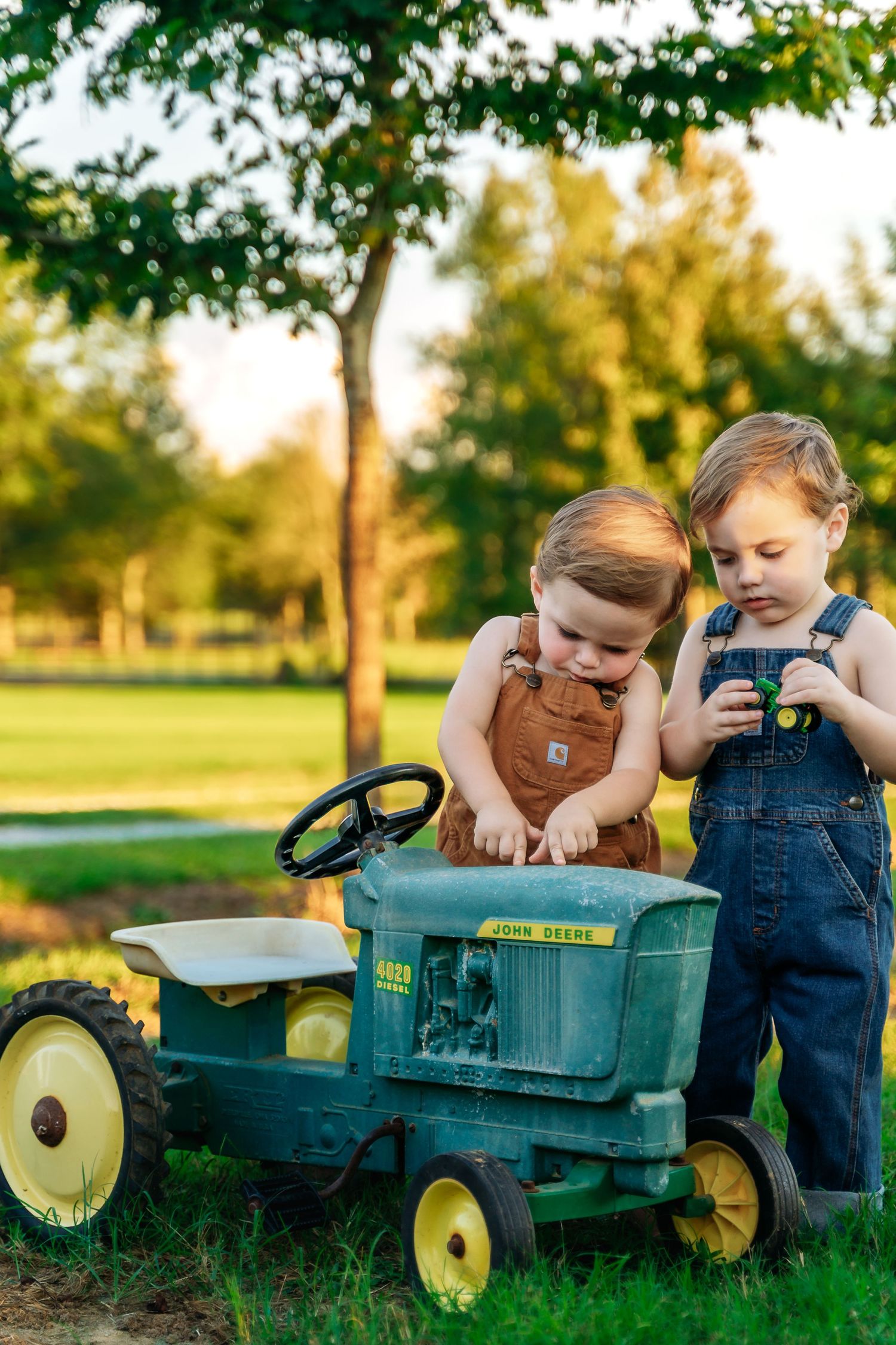 Two small children in overalls playing with a vintage green pedal tractor in a sun-drenched park setting.