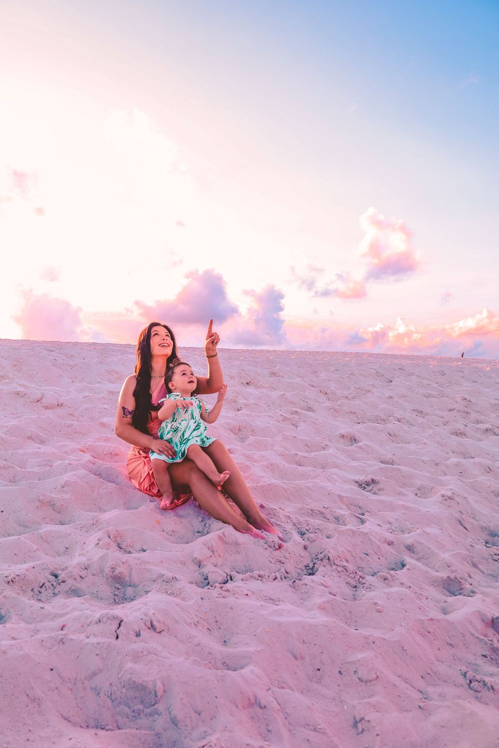 Two people sitting together on pink sandy beach at sunset with pastel colored sky in the background.