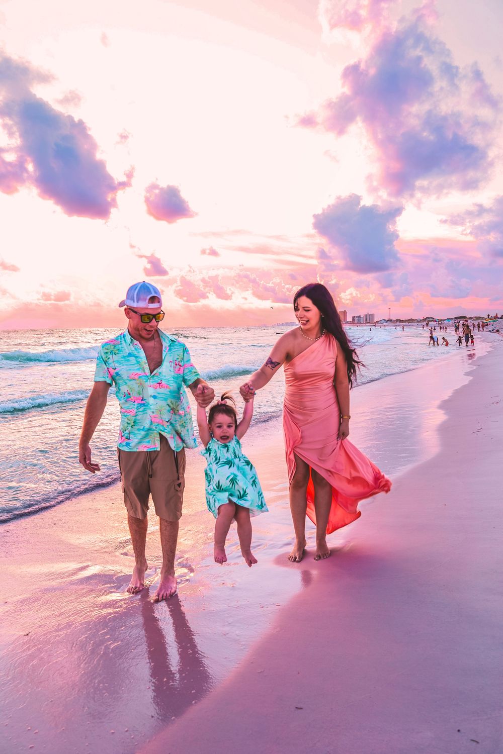 Family enjoying sunset walk along beach shoreline with colorful pink and purple sky reflecting on white sand.