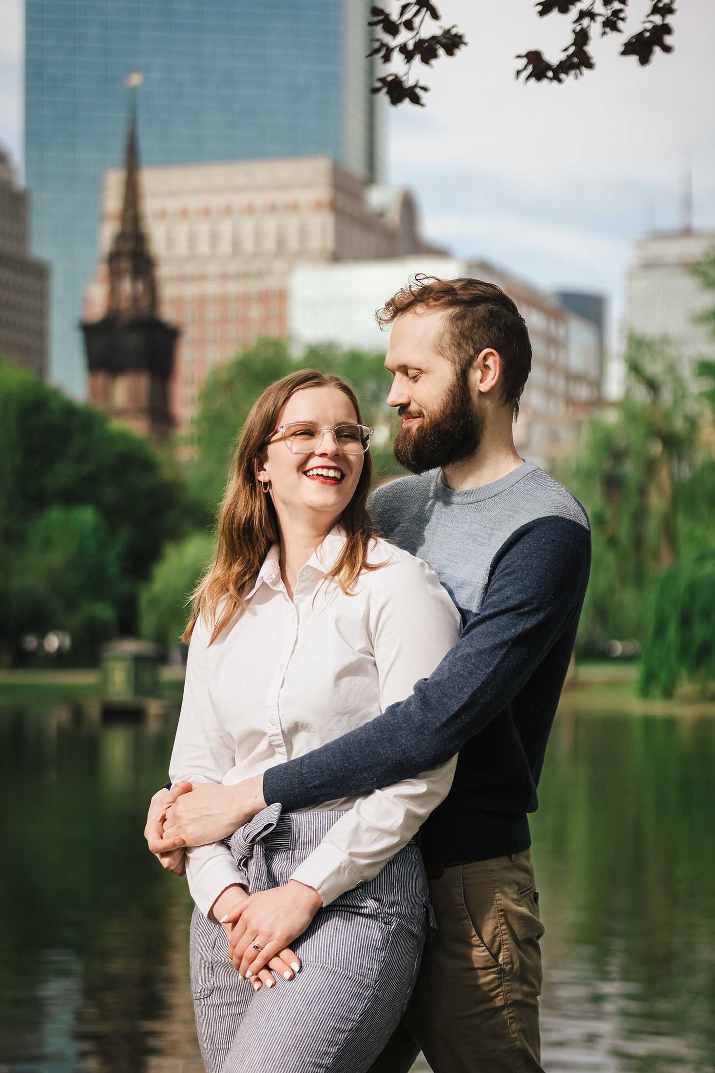 Romantic portrait of engaged couple with iconic Boston skyline and historic church spire in the background of Public Garden