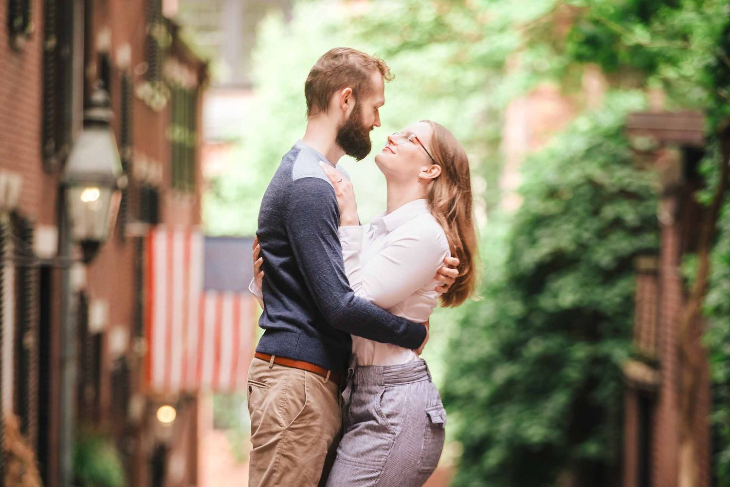 A close-up of a couple gazing into each other’s eyes while embracing on Acorn Street, with a softly blurred historic buildings in background
