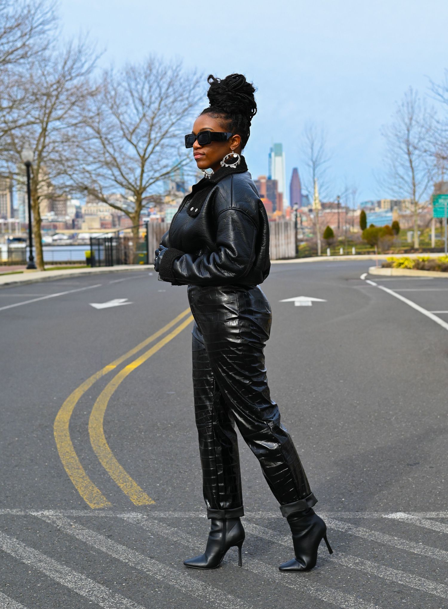 Stylish person wearing black leather outfit with heels poses on city street with urban skyline in background.