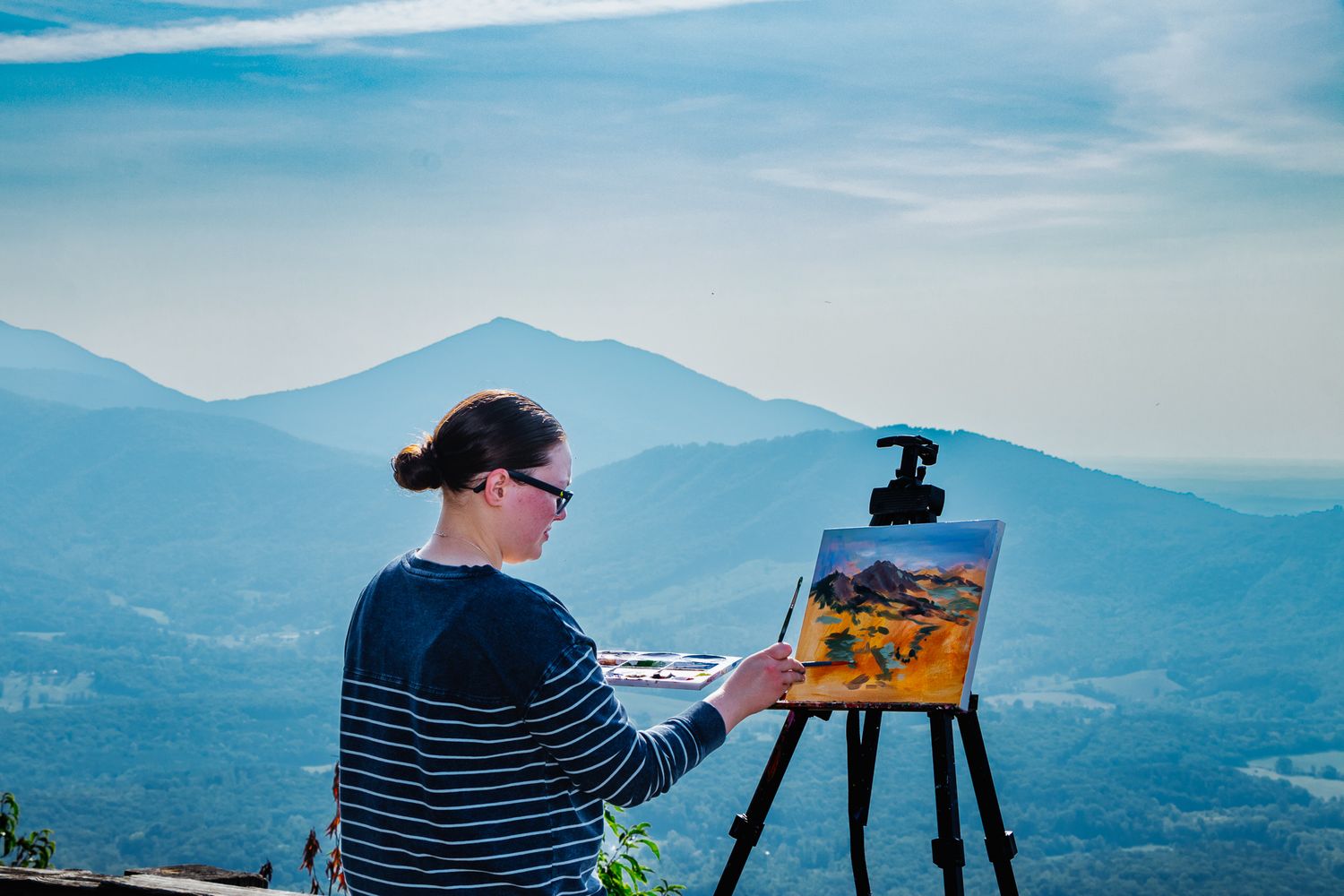 An artist painting a landscape on an easel, framed by the peaks of the Blue Ridge Mountains under a bright morning sky