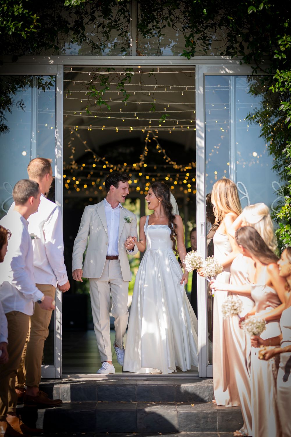 Wedding party lines the doorway as newlyweds exit under twinkling lights.