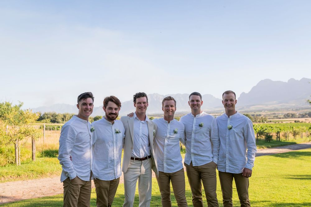 A group of groomsmen in light colored shirts pose together on a lawn with mountains in the background.