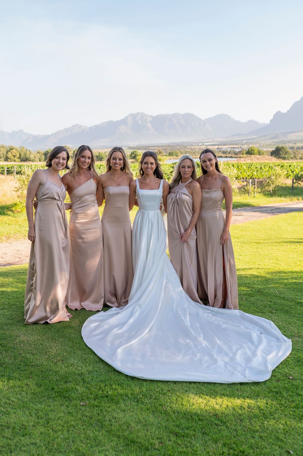 Bridesmaids in champagne dresses stand with bride in white gown against mountain backdrop at outdoor venue.