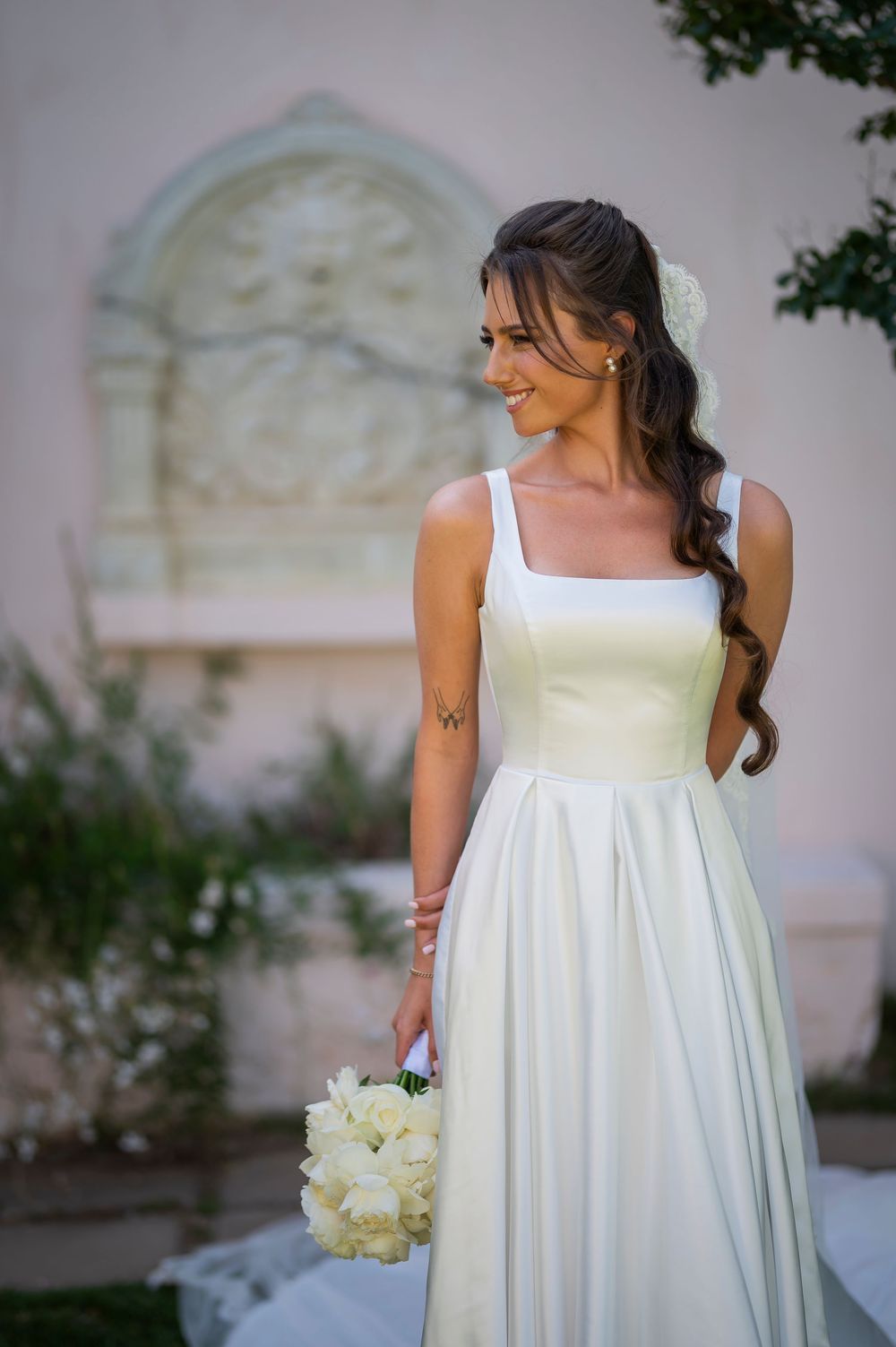 An elegant wedding dress with a square neckline and flowing skirt is shown in a garden setting.