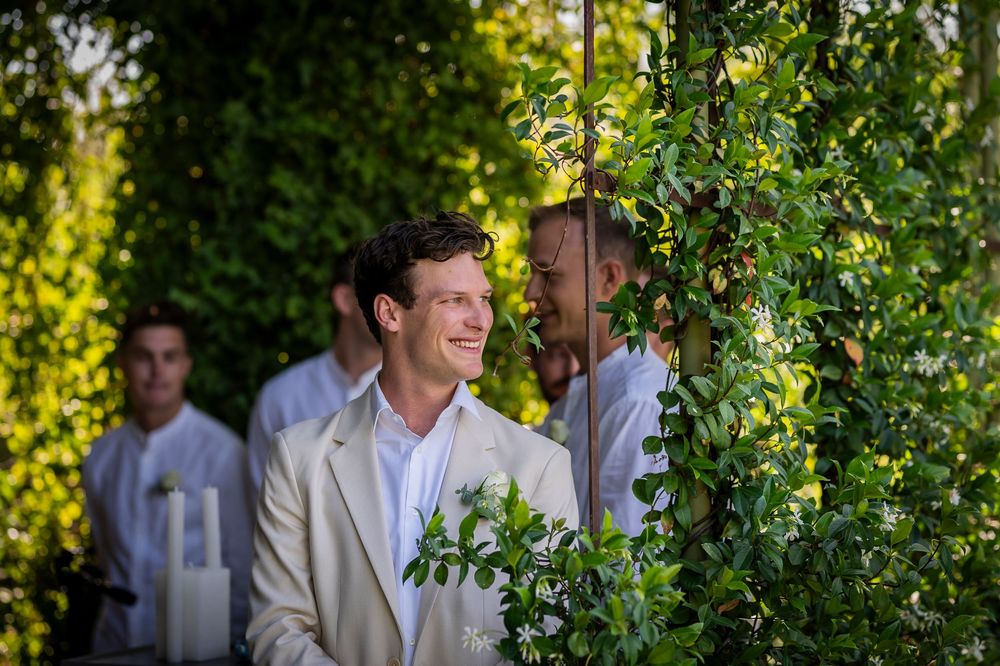 A guest in a light suit stands among green foliage at an outdoor wedding.