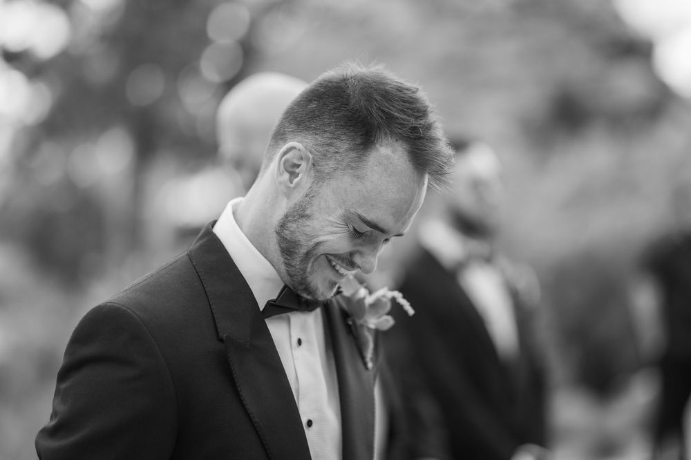 Black and white photo of a smiling groom in a tuxedo with bow tie at an outdoor wedding ceremony.