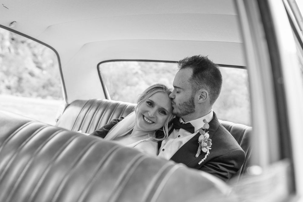 Black and white photo of a bride and groom sharing a romantic kiss in the backseat of a vintage car.