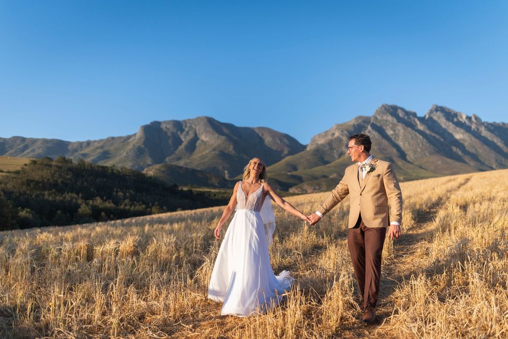 Bride and groom holding hands in golden field with stunning mountain backdrop under clear blue sky.
