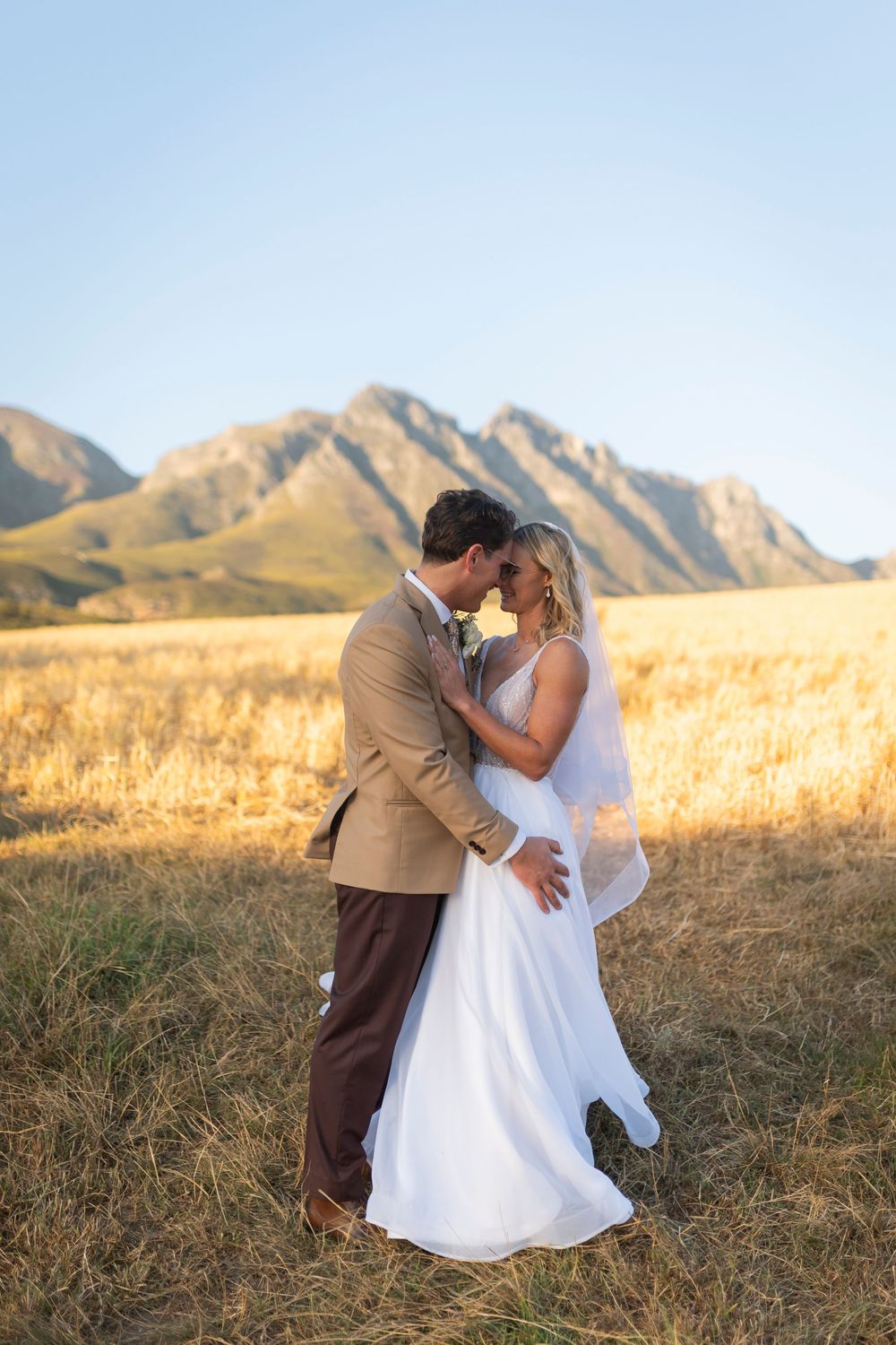 Bride and groom share romantic moment in golden field with mountain backdrop on sunny wedding day.
