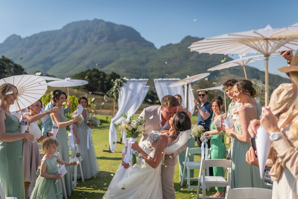 Bride and groom share a romantic dip kiss at outdoor wedding ceremony with mountain backdrop and guests holding parasols.