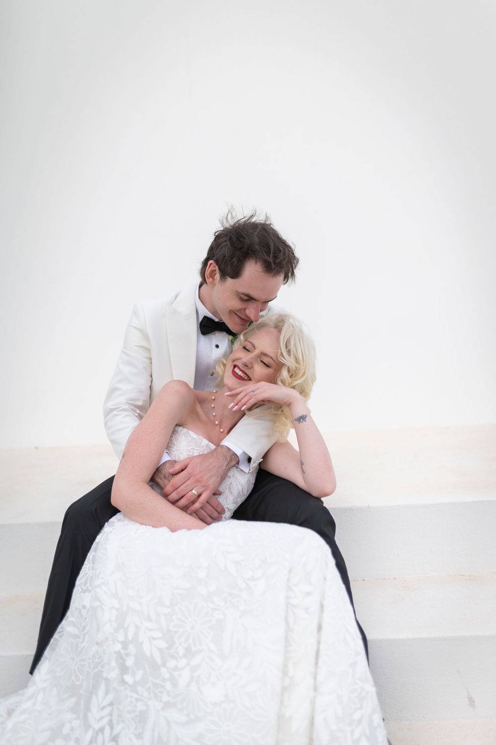 A joyful bride in a white lace gown sits with her groom in a white tuxedo jacket, sharing a tender embrace.