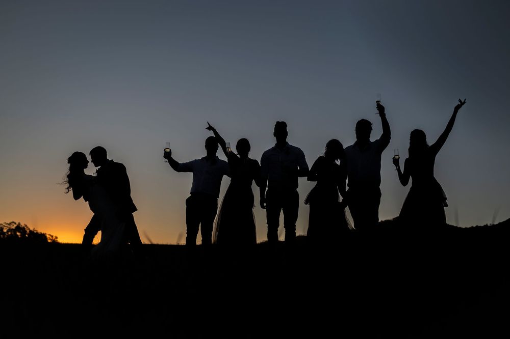 Wedding party silhouettes pose playfully against a dramatic sunset sky.