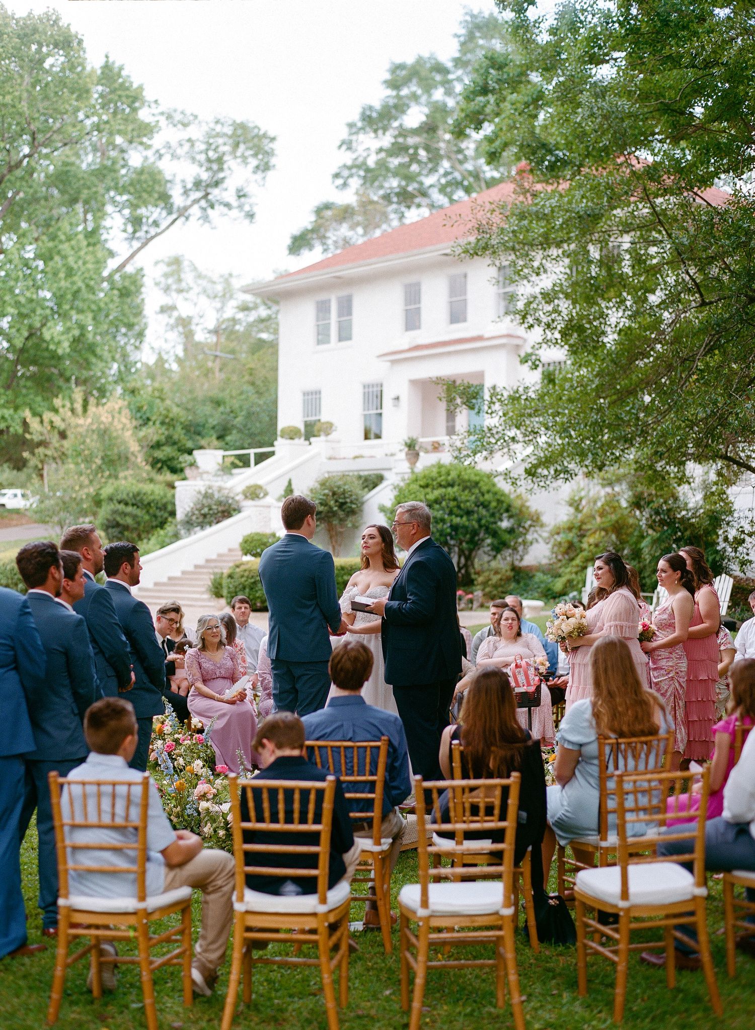 Garden wedding ceremony setup with white chairs and guests on a lawn in front of a colonial style house.