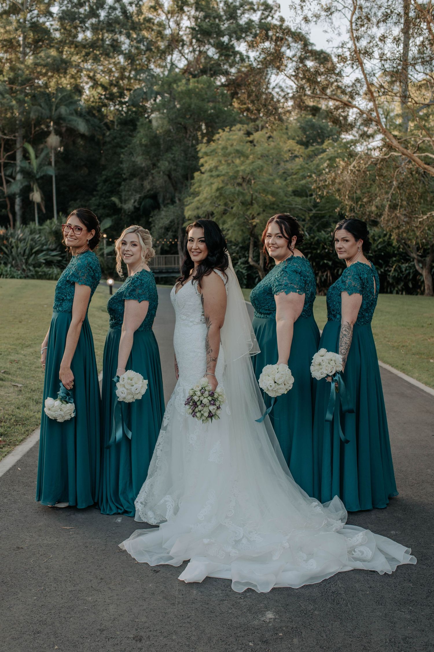 Bride in white gown poses with four bridesmaids wearing matching teal floor-length dresses holding white bouquets outdoors.